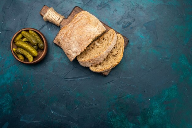 Вид сверху буханки хлеба с солеными огурцами на темно-синем столе.
