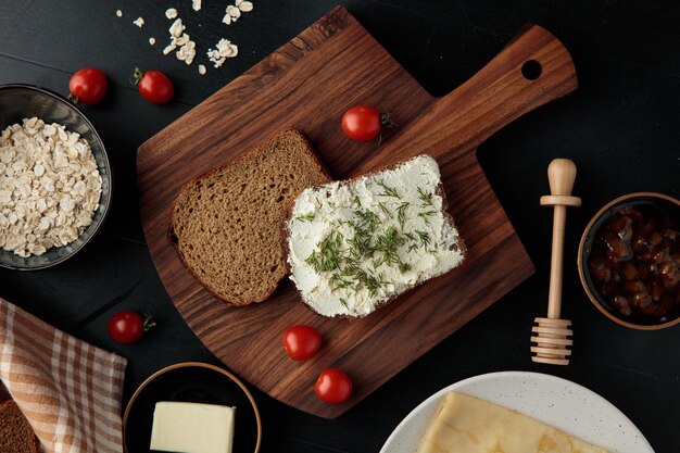 Top view of bread slices one smeared with cottage cheese with dill on it and tomatoes on cutting board and oats with jam and butter on black background