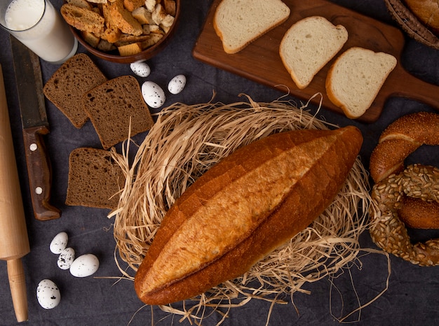 Top view of breads as baguette on straw rye and white breads bagel with milk eggs on maroon background