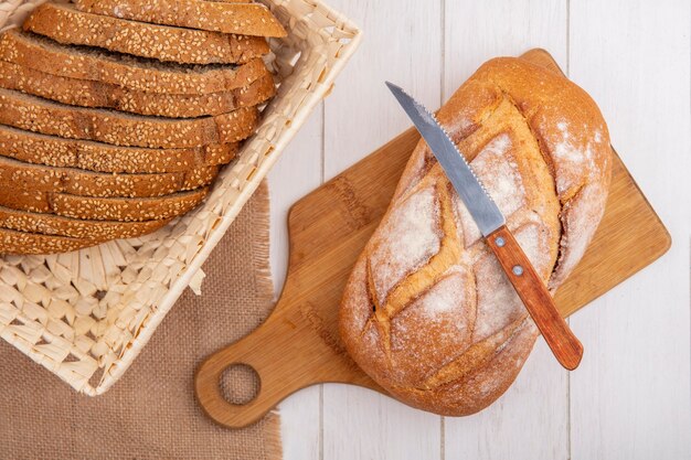 Top view of breads as brown sliced seeded cob in basket on sackcloth and crusty bread with knife on cutting board on wooden background