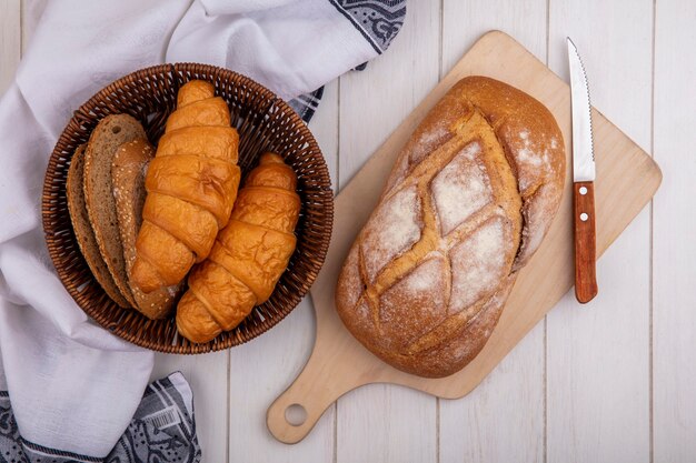 Вид сверху хлеба в виде круассана и кусочков коричневого початка с семенами в корзине на ткани и хрустящего хлеба на разделочной доске на деревянном фоне
