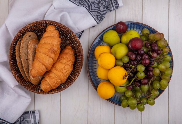 Top view of breads as croissant and seeded brown cob bread slices in basket on cloth and plate of grape nectacot pluot on wooden background