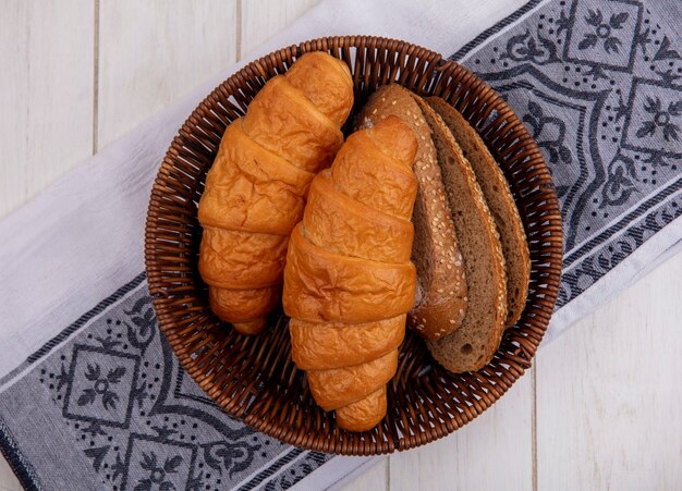 木製の背景の布の上のバスケットにクロワッサンと種をまく茶色の穂軸パンのスライスとしてパンの上面図