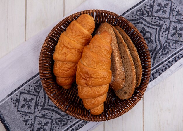 Top view of breads as croissant and seeded brown cob bread slices in basket on cloth on wooden background