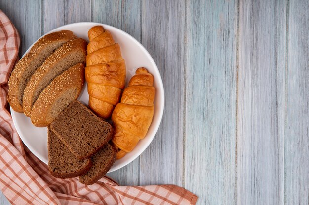 Top view of breads as croissant sliced rye and seeded brown cob in plate on plaid cloth on wooden background with copy space
