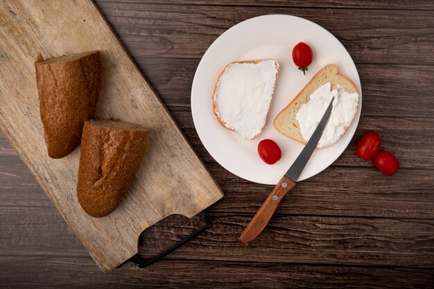 Top view of breads as cut in half baguette on cutting board and plate of sliced white bread with tomatoes and knife on wooden background