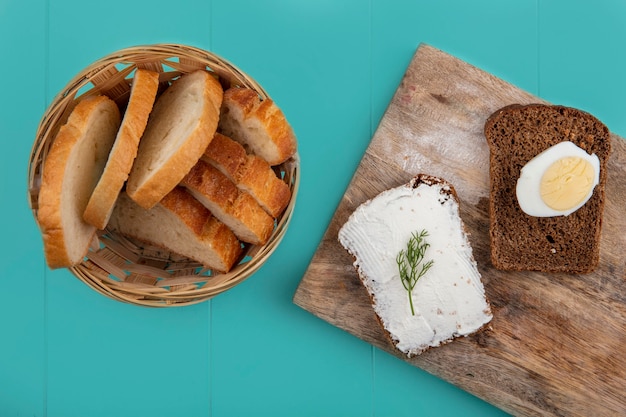 Free photo top view of breads as sliced baguette in basket and rye bread slice smeared with cheese and egg on cutting board on blue background