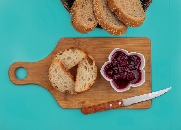 Top view of breads as sliced baguette seeded brown cob ones in basket and on cutting board with raspberry jam and knife on blue background