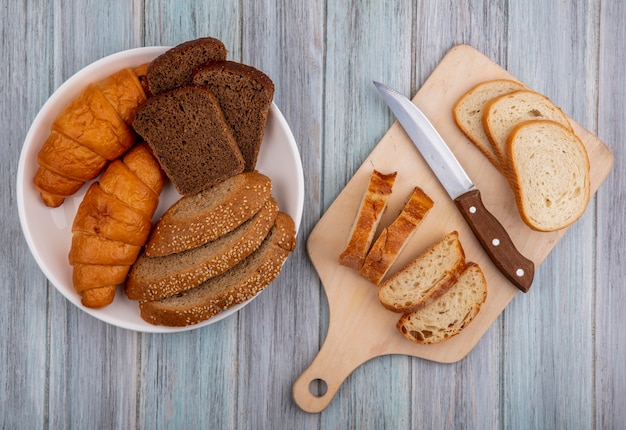 Top view of breads as sliced baguette with knife on cutting board and croissant rye and seeded brown cob in bowl on wooden background