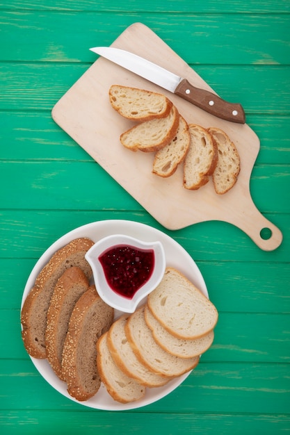 Top view of breads as sliced croissant with knife on cutting board and sliced seeded brown cob and baguette ones with raspberry jam on green background