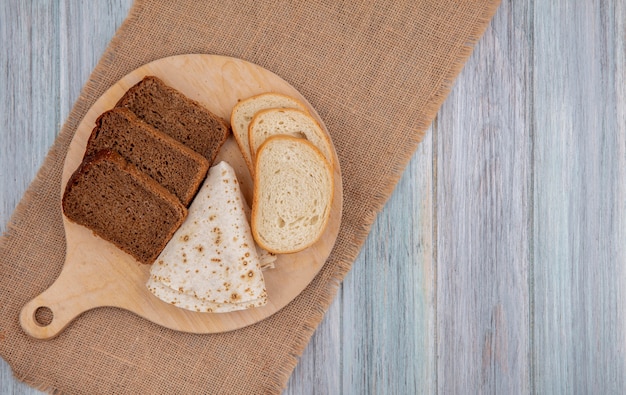 Top view of breads as sliced rye white ones and flatbread on cutting board on sackcloth on wooden background with copy space