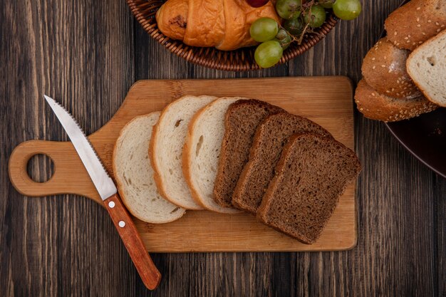 Top view of breads as sliced rye and white ones with knife on cutting board and basket of croissant grape with bowl of seeded brown cob slices on wooden background