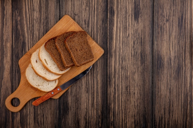 Top view of breads as sliced rye and white ones with knife on cutting board on wooden background with copy space