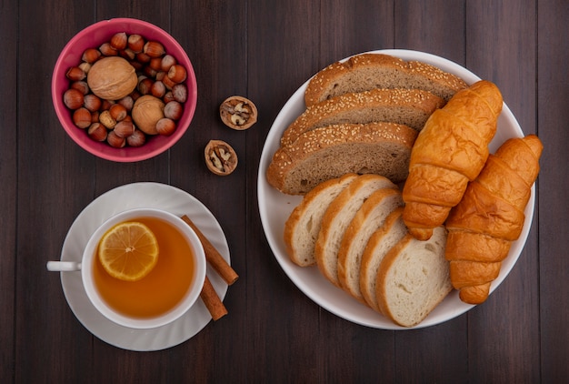 Top view of breads as sliced seeded brown cob baguette and croissants in plate and bowl of nuts walnuts and cup of hot toddy with cinnamon on saucer on wooden background