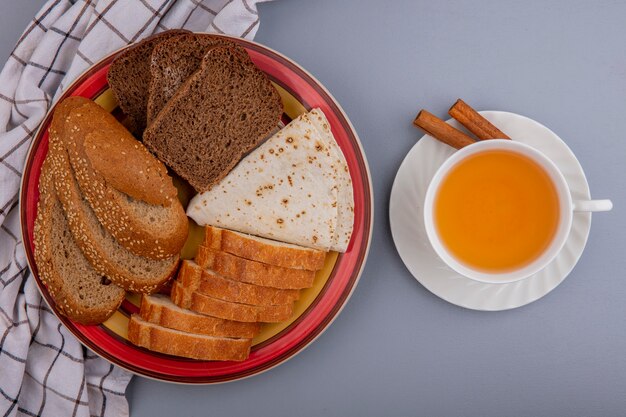 Top view of breads as sliced seeded brown cob rye baguette and flatbread in plate on plaid cloth and cup of hot toddy with cinnamon on saucer on gray background