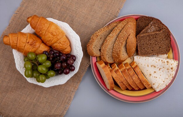 Top view of breads as sliced seeded brown cob rye baguette and flatbread in plate on plaid cloth with plate of croissants grapes on sackcloth on gray background