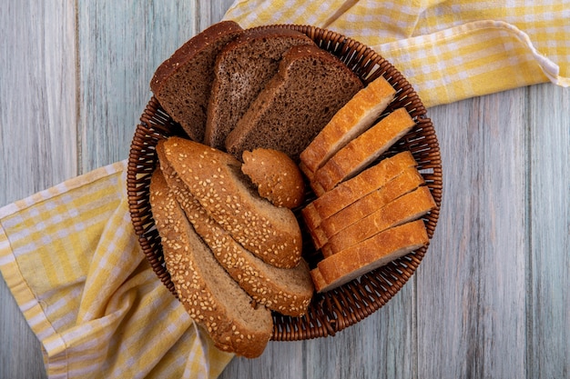 Top view of breads as sliced seeded brown cob rye and crusty ones in basket on plaid cloth on wooden background