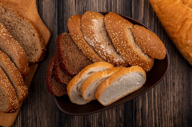 Top view of breads as sliced seeded brown cob rye and white ones in bowl and on cutting board on wooden background