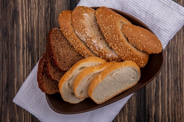 Вид сверху на хлеб в виде нарезанных семенами коричневой ржи и белых хлебов в миске на белой ткани на деревянном фоне