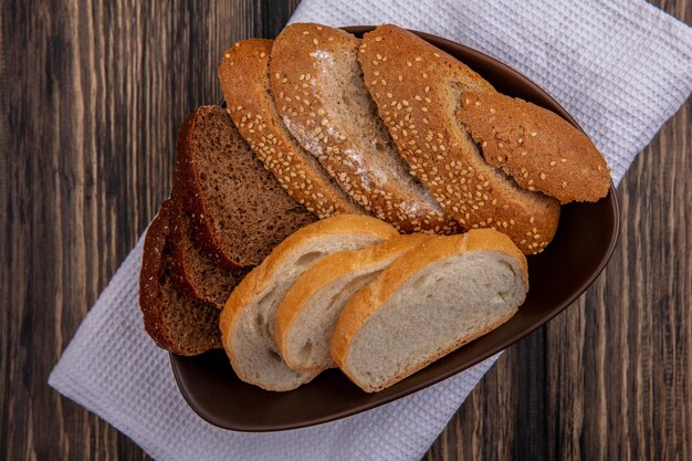 Top view of breads as sliced seeded brown cob rye and white ones in bowl on white cloth on wooden background
