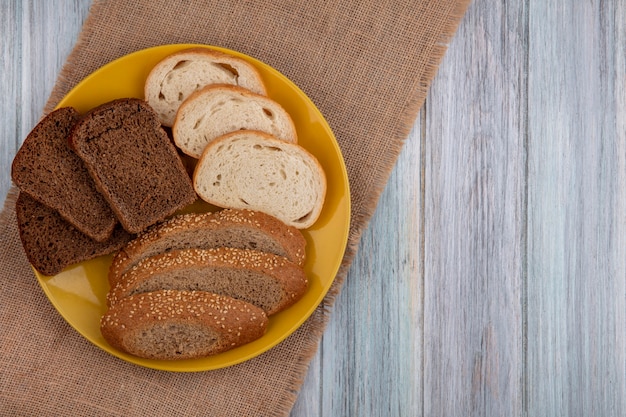 Вид сверху на хлеб в виде нарезанных семенами коричневого початка ржи белые в тарелке на вретище на деревянном фоне с копией пространства