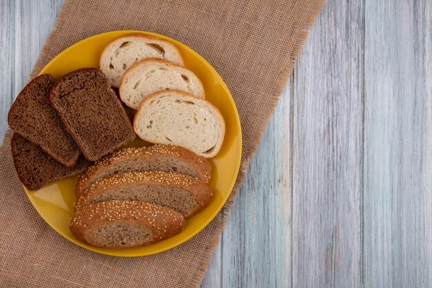 Top view of breads as sliced seeded brown cob rye white ones in plate on sackcloth on wooden background with copy space