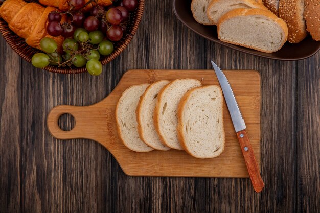 Top view of breads as sliced seeded brown cob and white ones in bowl and on cutting board with knife and basket of croissant grape on wooden background