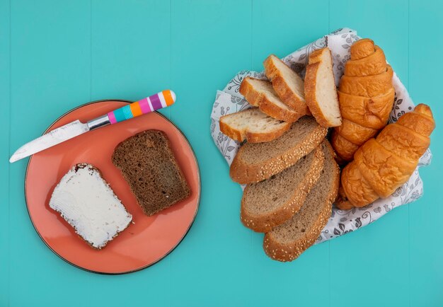 Вид сверху хлеба в виде нарезанного семенами багета из початков и круассана в миске и ржаного хлеба, намазанного сыром с ножом в тарелке на синем фоне