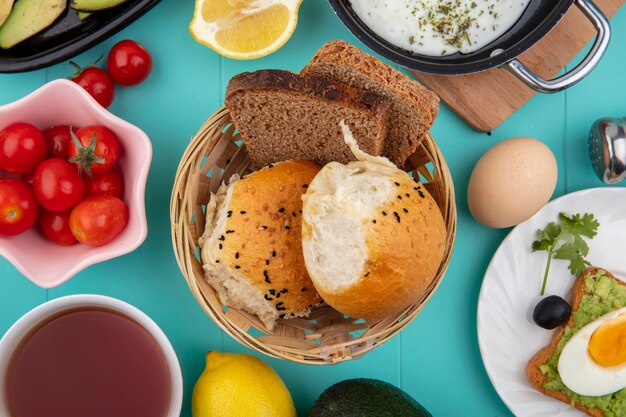 Вид сверху хлеба на ведре с помидорами, жареным яйцом с лимоном и яйцом на сковороде на деревянной кухонной доске на синем