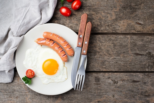 Top view of breakfast egg and sausages on plate with tomatoes and cutlery