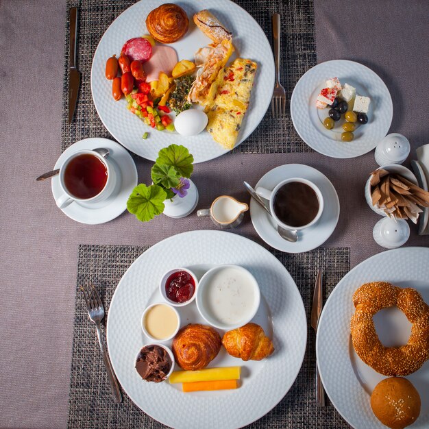 Top view breakfast eggs, olives, omelette, croissant in plates and cup of tea on table