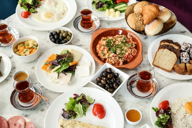 Top view breakfast set scrambled eggs with tomatoes a variety of cheeses vegetables olives honey with tea and bread on the table