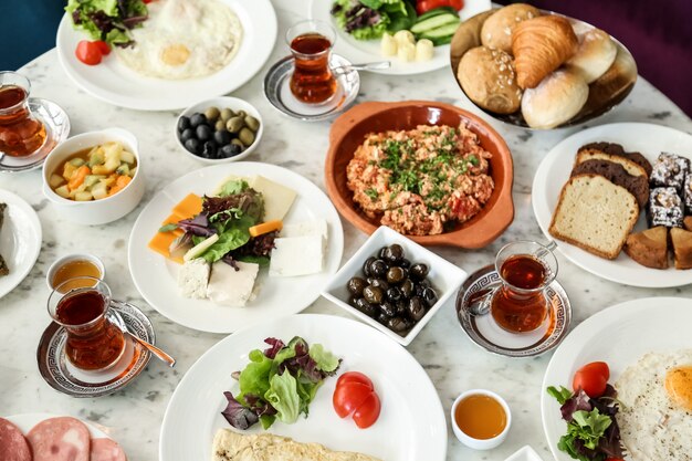 Вид сверху на завтрак - яичница-болтунья с помидорами, различными сырами, овощами, оливками, медом, чаем и хлебом на столе.