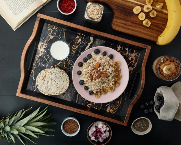 Top view of breakfast set with banana blackthorn almond flax oatmeal and crispbreads with glass of milk in tray and banana walnut smoothie with ingredients around for making breakfast