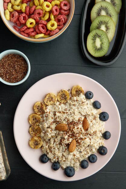 Вид сверху на завтрак с банановой терновой льняной миндальной овсянкой в тарелке и миской хлопьев с нарезанным киви и семенами льна на черном фоне
