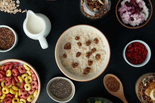 Вид сверху на набор для завтрака с миской грецкого ореха, овсяной кашей, миской хлопьев с молоком, красной смородиной, семенами льна, чиа, творогом, грецким орехом на черном фоне