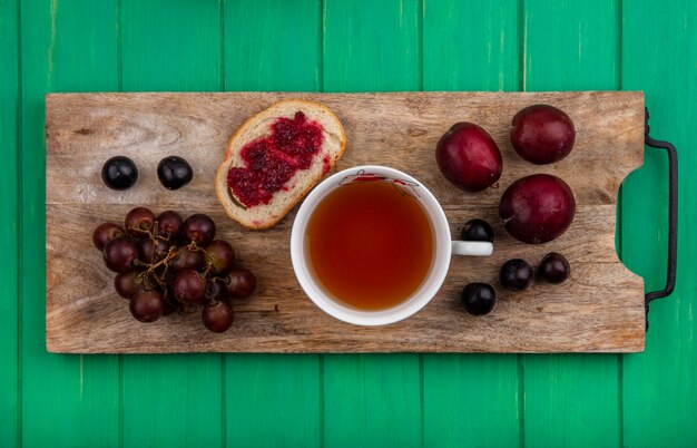 Top view of breakfast set with bread slice smeared with raspberry jam grape cup of tea and pluots with sloe berries on cutting board on green background