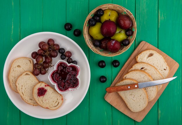 Top view of breakfast set with bread slices raspberry jam and grape in plate and bread slices with knife on cutting board with basket of pluots on green background