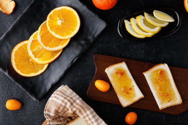 Top view of breakfast set with bread slices smeared with jam on cutting board and orange slices in plate with lemon slices on black background