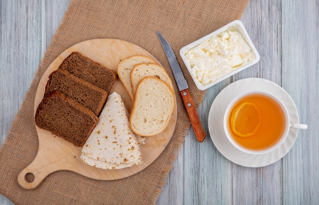 Top view of breakfast set with breads as sliced rye white ones and flatbread on cutting board with knife and clotted cream on sackcloth and cup of hot toddy on wooden background