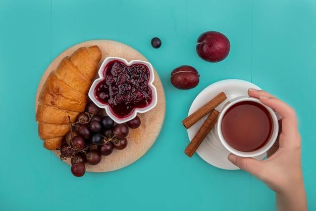 Top view of breakfast set with croissant and raspberry jam grape on cutting board and female hand holding cup of tea with cinnamon and pluots on blue background