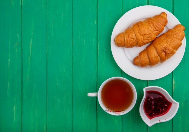 Top view of breakfast set with croissants in plate cup of tea raspberry jam in bowl on green background with copy space