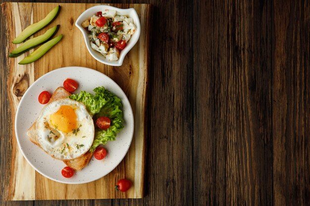 Top view of breakfast set with fried egg lettuce tomatoes on dried bread slice in plate and avocado slices with vegetable salad on cutting board on wooden background with copy space