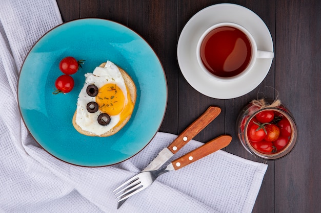 Free photo top view of breakfast set with fried egg olive and tomatoes in plate with fork and knife on cloth and bowl of tomato with cup of tea on wood