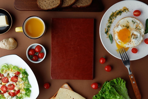 Vista dall'alto del set per la colazione con uovo fritto con pomodoro e aneto e insalata di verdure con pomodoro, lattuga all'uovo con forchetta, succo d'arancia, fette di pane e tagliere al centro