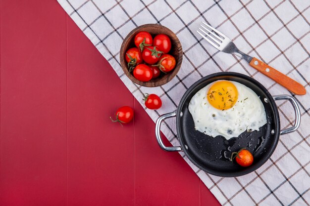 Top view of breakfast set with pan of fried egg and bowl of tomato fork on plaid cloth on red with copy space