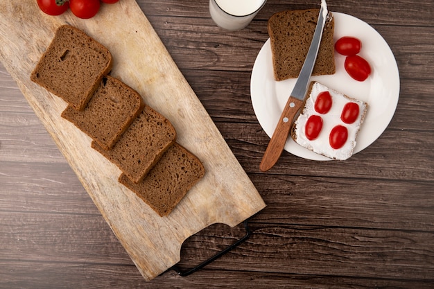 Top view of breakfast set with sliced rye bread smeared with cottage cheese and tomatoes with knife in plate on wooden background