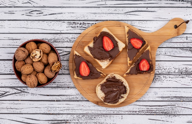 Top view of breakfast toasts with chocolate and strawberry on cutting board and walnuts in bowl on white wooden surface horizontal