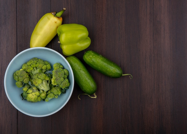 Top view broccoli in bowl with bell peppers and cucumbers on wooden background