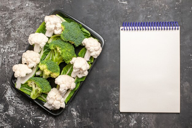 Top view broccoli and cauliflower on black rectangular plate a notebook on dark background